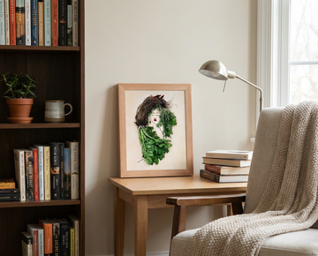 The Celery Woman botanical wall art in an oak frame, displayed on a wood desk in a calm reading corner with a bookshelf.